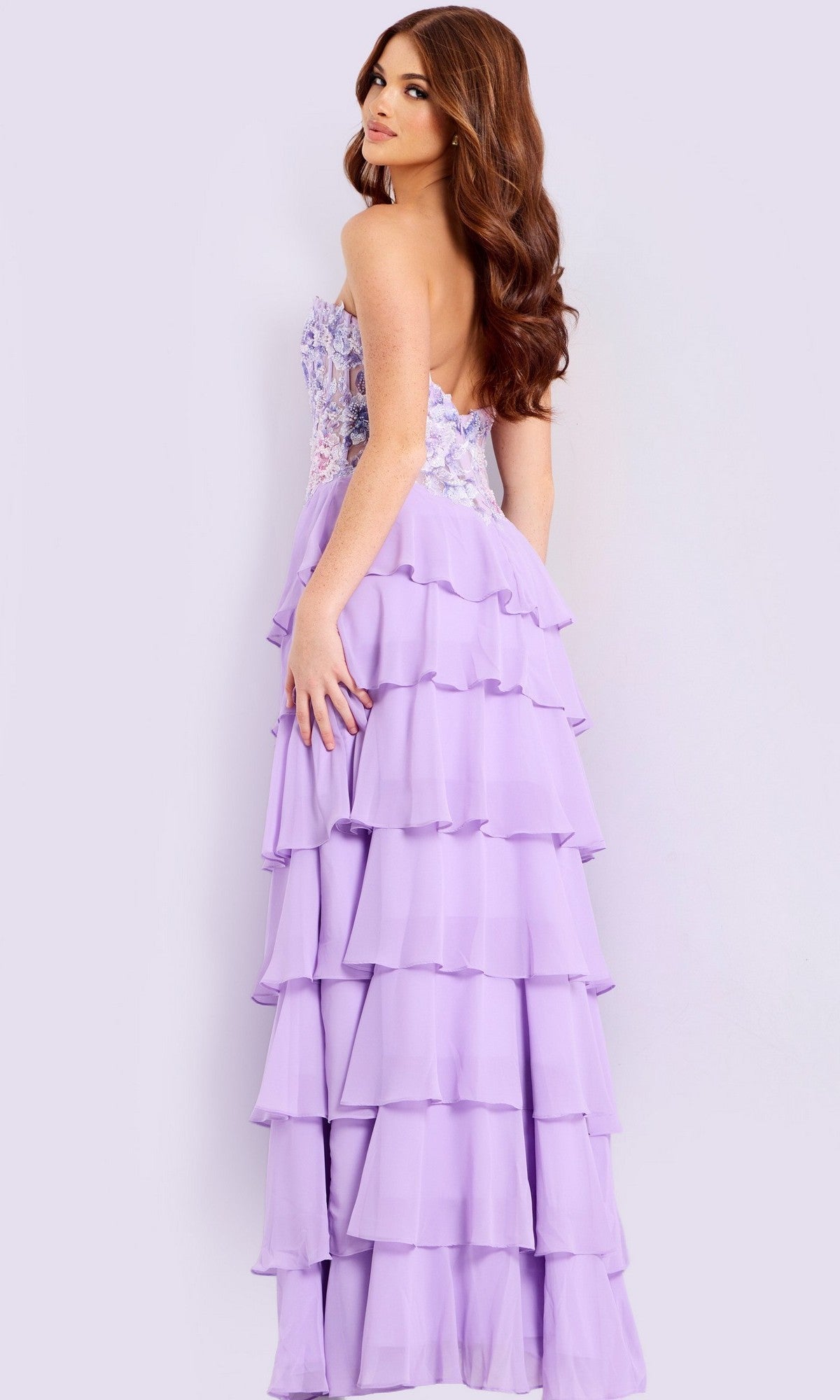 Woman wearing the Jovani 49628 long prom dress, featuring a strapless bodice with floral embroidery and a lavender ruffled layered chiffon skirt, standing and looking over her shoulder.